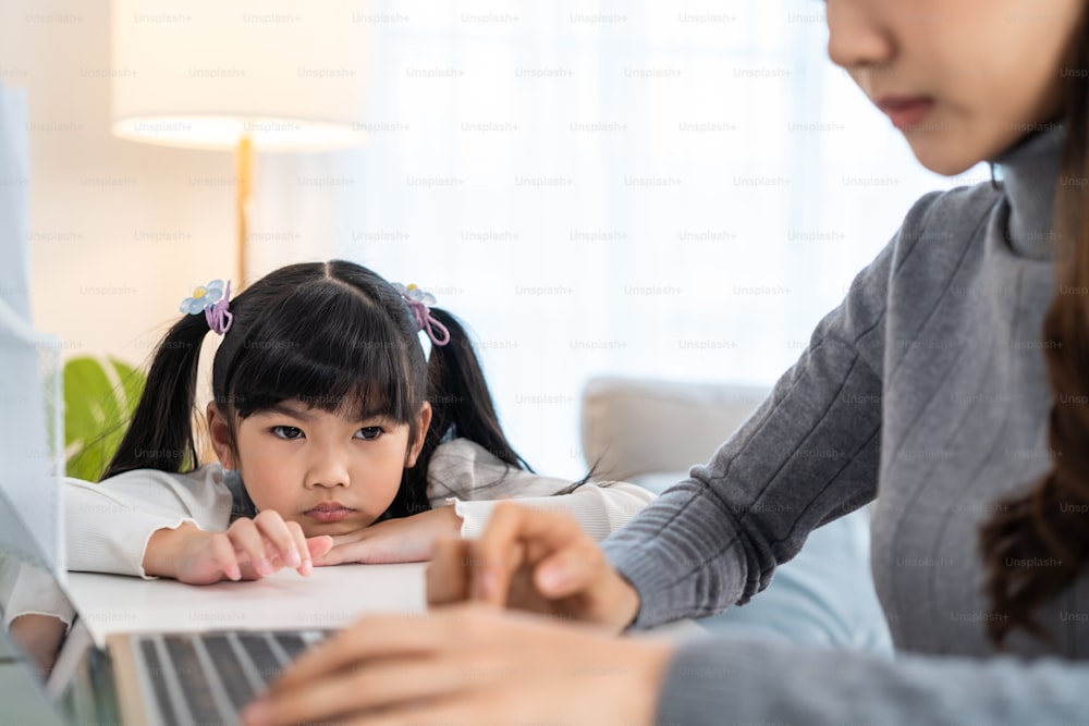 Asian young kid daughter feeling sad and need to play with busy mother. Attractive beautiful mom use laptop computer working from home during new normal and have no time for little child girl in house