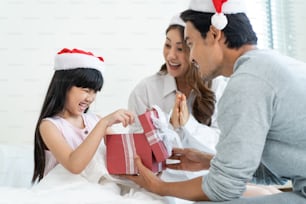 Asian lovely young little daughter open present gift box from parents. Happy family, little kid feel happy and excited for surprise from mother and father ready to celebrate Christmas together at home