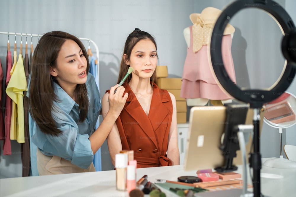 Asian two women vlogger talk to camera to review the cosmetic at home. Beautiful Beauty blogger Influencer woman friends couple live streaming and showing makeup brush to followers in office store.