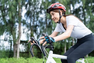 公共の公園で夕方に自転車に乗っているアジアの若いスポーツの女性。スポーツウェアを着た美しいアスリートのフィット感としっかりした女の子はヘルメットを着用し、彼女のヘルスケアの健康と新鮮なサイクリングトレーニングで運動します。