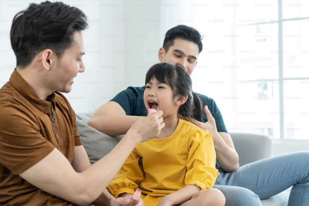 Asian attractive LGBTQ gay family put lipstick on young girl kid mouth. Handsome male couple wearing makeup and combing little adorable child daughter's hair in living room, enjoy parenting at home.