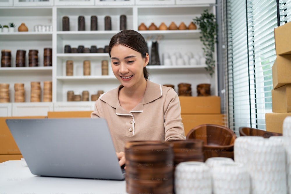 Mulher bonita asiática vender produto de vaso e verificar a ordem de venda no laptop. Jovem empresária atraente é feliz e sorrir depois de olhar para o financeiro dos negócios. Conceito de compra e compra remota.