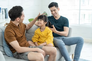 Asian attractive LGBTQ gay family spend time with girl kid daughter. Handsome male couple sit on sofa in living room, take care and comb little adorable child's hair enjoy parenting activity at home.
