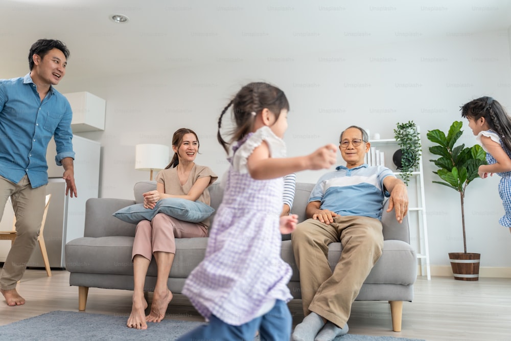 Asian family spending time together on holiday in living room at home. Attractive happy parents, father play with young little two kid girl daughter run around sofa in house. Activity relationship.