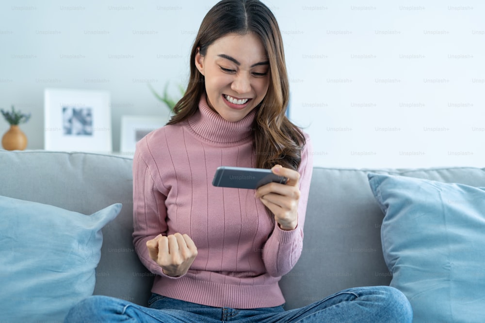 Asian young beautiful woman playing mobile game on smartphone at home. Attractive casual girl feel happy and relax, sit on sofa having fun touching screen on phone to play video enjoy victory in house