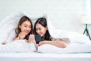 Asian attractive lesbian couple use mobile phone watch movie on bed. Beautiful sibling in pajamas lying down with blanket in bedroom together, use smartphone look at video online in morning in house.