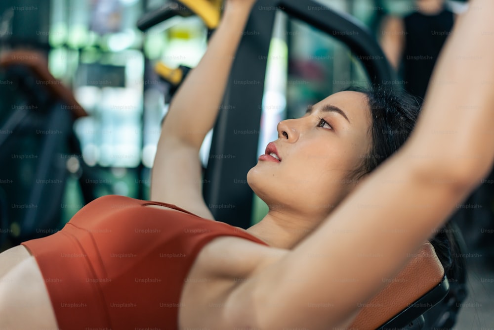 Asian sportswoman slowly lift up weight machine or barbell in stadium. Attractive young athlete female workout exercise use weight equipment tool for health care and maintain muscle in fitness club.