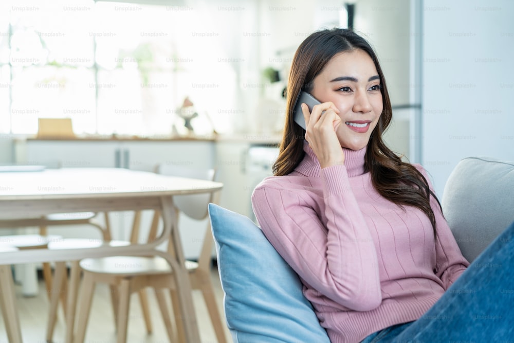 Asian beautiful woman sit on sofa and talk on mobile phone in house. Happy attractive young girl  spend leisure time at home, feel relax and enjoy communicate and discuss on smartphone in living room.