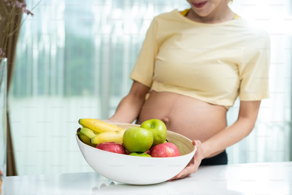 Close up of Asian woman pregnant standing and holding mix fruit bowl. Attractive beautiful young female pregnancy stand and touching baby in the womb, choose healthy food or vegetarian food in kitchen