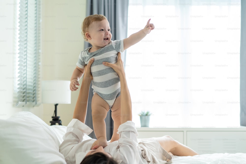 Caucasian loving mom play with cute baby boy child on bed in bedroom. Happy family, attractive beautiful caring young mother lifting and talking with toddler infant son in the early morning in house.