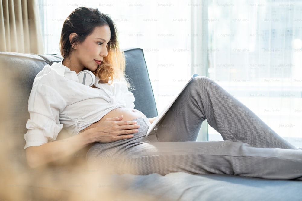 Asian young woman pregnant sitting and reading book with smile on sofa. Attractive female pregnancy sit on couch looking guidebook preparing for baby in womb, with happiness and smiling in living room
