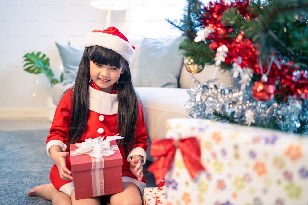 Asian young little daughter open present gift box under Christmas tree. Happy family, little kid feel happy and excited for surprise from mother and father ready to celebrate Xmas together in house.