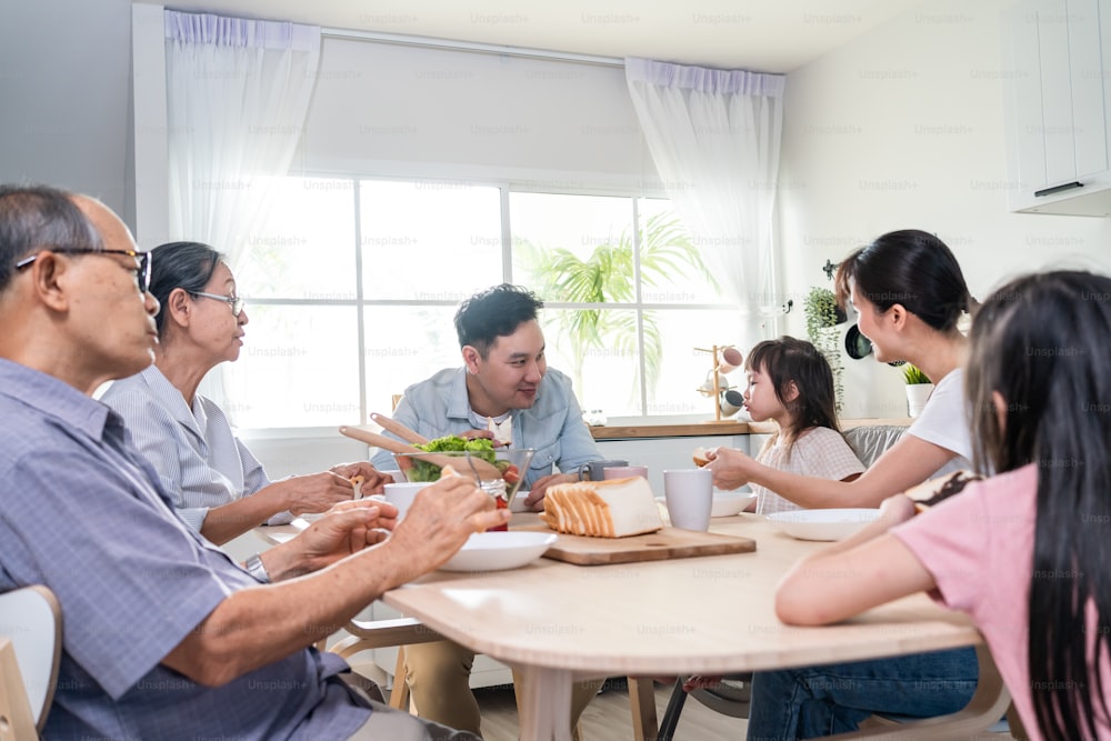 La gran familia asiática feliz almuerza en la mesa de comer juntos en la casa. El abuelo mayor, la pareja joven y las hijas pequeñas se sienten felices, disfrutan comiendo pan en casa. Concepto de relación de actividad