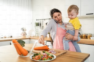 Caucasian busy mother doing housework with baby boy toddler in kitchen. Beautiful single mom use phone call for work and cook foods prepare breakfast for little kid son in house. Family-housekeeping.
