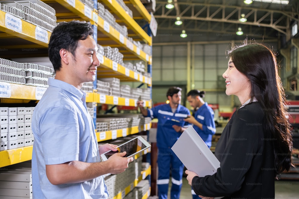 アジアのビジネスウーマンは、パートナーと一緒に工業製造工場で働いています。フォーマルウェアを着た魅力的な業界の工場エンジニアの人々は、製造所の倉庫で注文と製品を幸せに処理します。