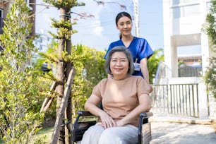 Asian happy senior woman patient sitting on wheelchair at green park. Caregiver nurse push and take elderly older female handicap outdoor for physical therapy hearth care activity at nursing home.