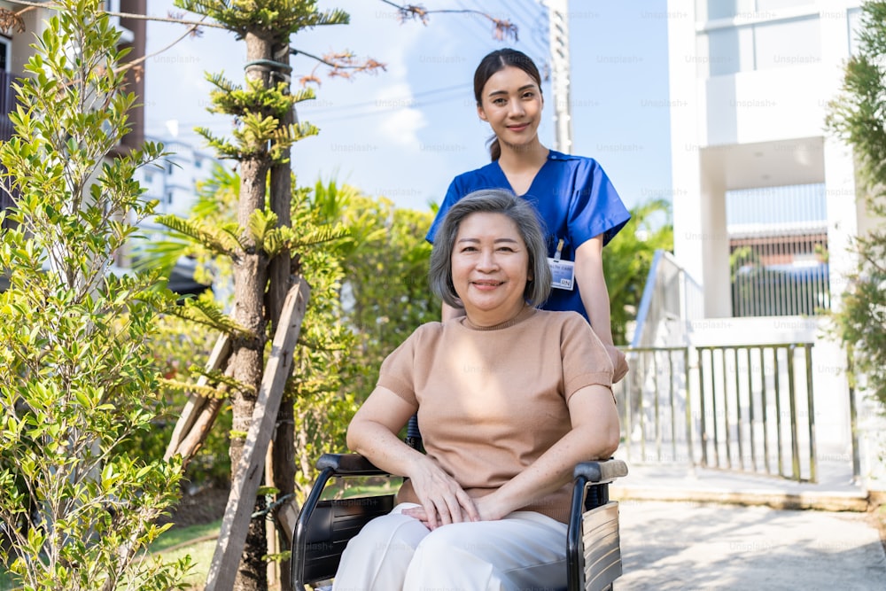 Paciente sênior asiático feliz sentado na cadeira de rodas no parque verde. Enfermeira cuidadora empurra e leva idosos idosos com deficiência feminina idosa ao ar livre para atividade de cuidados de lareira de fisioterapia em casa de repouso.