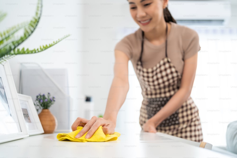 Femme de ménage asiatique qui nettoie dans le salon à la maison. Belle fille femme au foyer femme de ménage nettoyeur se sentir heureux et essuyer la table de travail sale et désordonnée pour le ménage, les tâches ménagères ou les tâches ménagères.