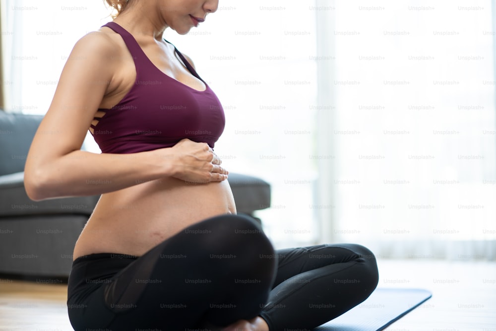 Giovane bella donna asiatica incinta che fa allenamento di Yoga Pilates a casa. Attraente gravidanza femminile in abbigliamento sportivo sentirsi felici e rilassarsi, seguire le istruzioni di esercizio dal trainer online in soggiorno.