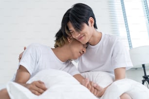 Portrait of Asian handsome gay couple sit on bed in bedroom at home. Attractive romantic male lgbt couple hugging each other, feeling happy to spend time together and smile looking at camera in house.