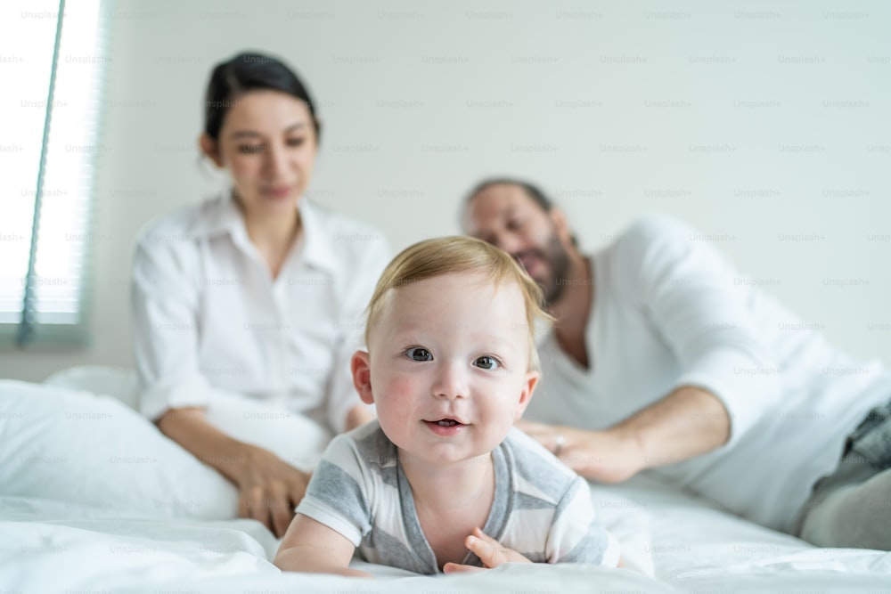 Caucasian parents playing with cute baby boy child on bed in bedroom. Happy family, attractive beautiful caring young mother and father feel happy look at toddler kid son crawling in morning in house.
