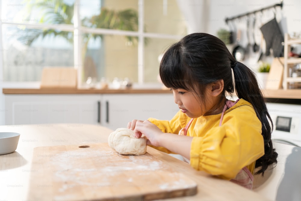 Portrait d’une jeune fille asiatique faisant de la boulangerie maison dans la cuisine. Adorable petit enfant assis sur une table se sentant heureux et appréciant d’apprendre à cuisiner des aliments ou à cuire de la pâte à levure avec les mains à la maison