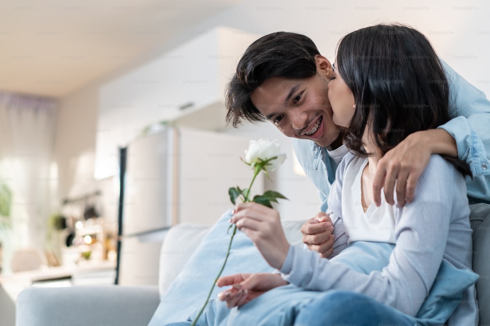 Asian young man surprise and give rose flower to beautiful girlfriend. Attractive romantic new marriage couple male and woman spend time together in living room at home, celebrate anniversary in house