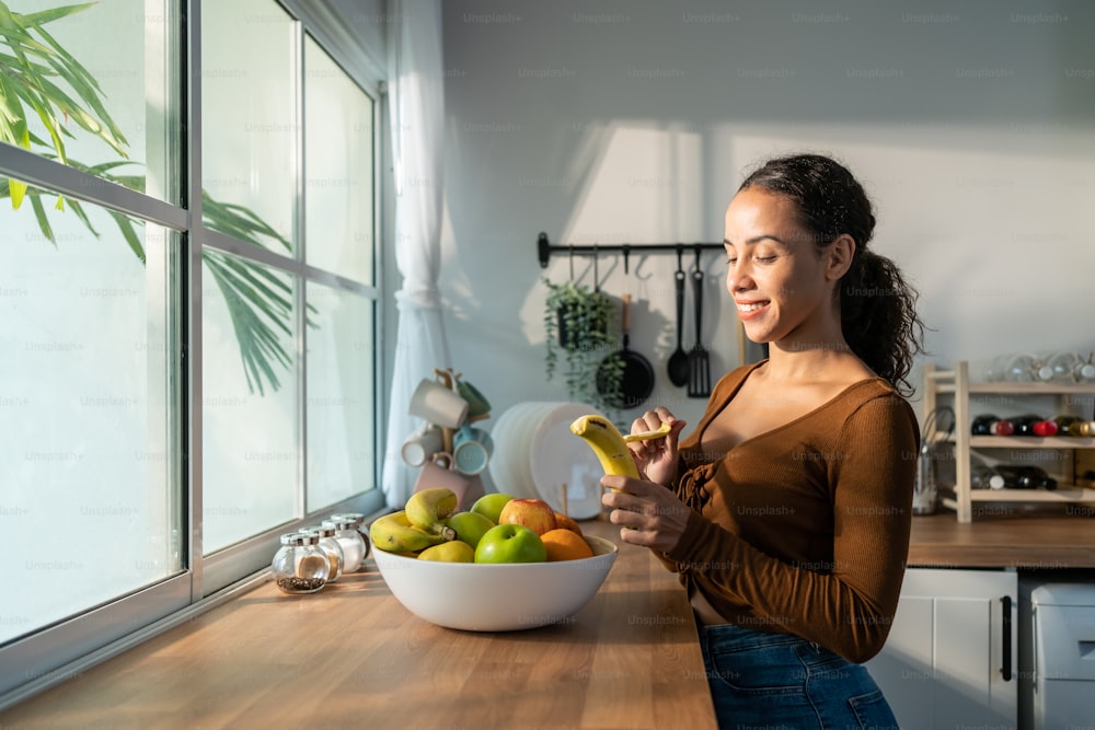 Junge attraktive Latino-Frau isst Früchte auf dem Tisch in der Küche zu Hause. Schöner aktiver weiblicher Stand mit glücklichem Gesicht, genießen Sie das Essen von Bananen, gesunde Lebensmittel für das Gesundheitswesen Wellness, um Gewicht im Haus zu verlieren