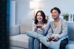 Asian young man and woman couple enjoy playing game together in house. Attractive funny male and female friend feeling happy sit on sofa in living room, spend time together play joystick game at home.