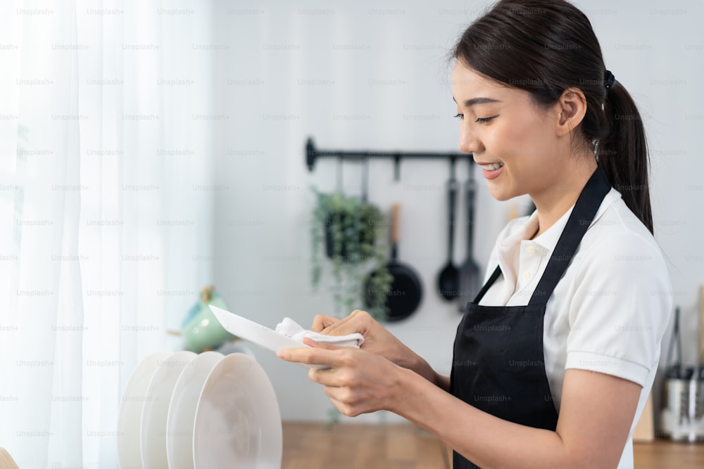 Servizio di pulizia attivo asiatico donna che pulisce in cucina a casa. La bella ragazza delle pulizie si sente felice e pulisce i piatti dopo il lavaggio per le faccende domestiche o le faccende domestiche.