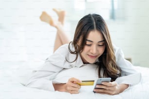Asian attractive woman enjoy shopping online on bed at home in morning. Casual female lying in bedroom, holding credit card to make e bank online payment after use phone purchase in web store in house