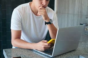 Thoughtful man freelancing with a laptop using a smart phone while sitting in the living room