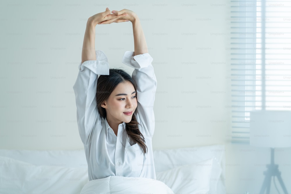 Asian beautiful girl in pajamas wake up in the morning with happiness. Attractive young woman smiling, feel happy and relax then stretching body after getting up from sleep on bed in bedroom at home.