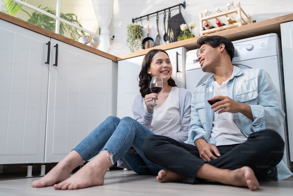 アジアの若い新婚カップルは、自宅のキッチンで一緒に時間を過ごします。魅力的なロマンチックな男性と女性は床に座って、家族関係の生活と新しい家への引っ越しを祝うためにワインを飲みます。