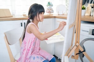 Asian young happy kid girl coloring on painting board in living room. Little adorable child learn how to draw art picture with watercolor paint and brush enjoy creativity activity on holiday at home.