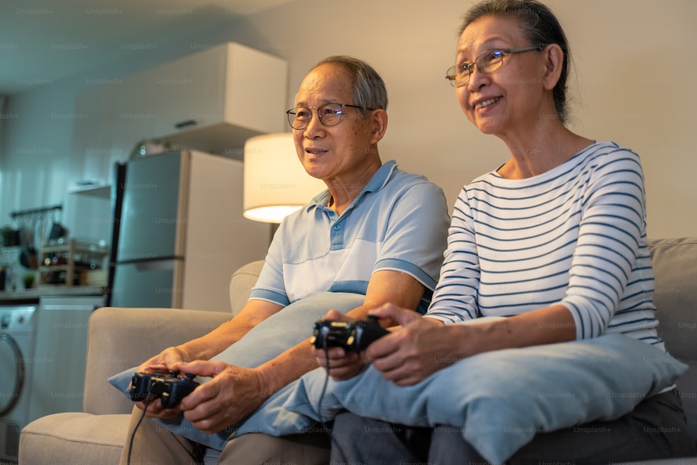 아시아 할아버지와 할머니는 집에서 함께 비디오 게임을 하고 있습니다. 매력적인 노인 노부부는 집에서 거실에서 웃는 얼굴로 흥분과 재미를 느끼며 게임 컨트롤러를 들고 앉아 있다