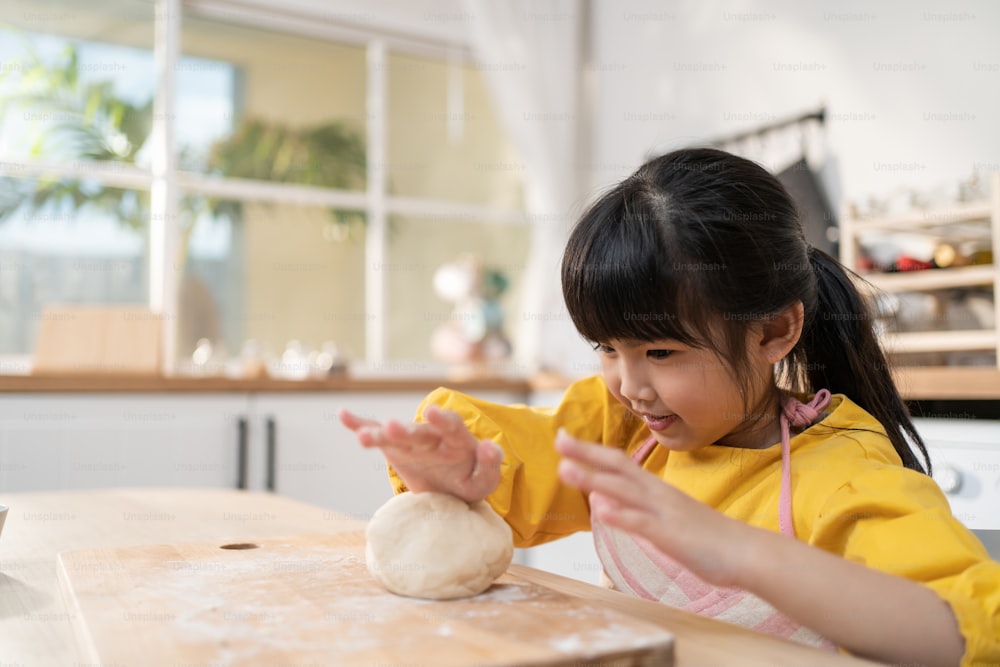 Porträt eines asiatischen jungen Mädchens, das hausgemachte Bäckerei in der Küche macht. Entzückendes kleines Kind, das auf dem Tisch sitzt und sich glücklich fühlt und genießt, zu lernen, wie man Lebensmittel kocht oder Hefeteig backt mit den Händen zu Hause