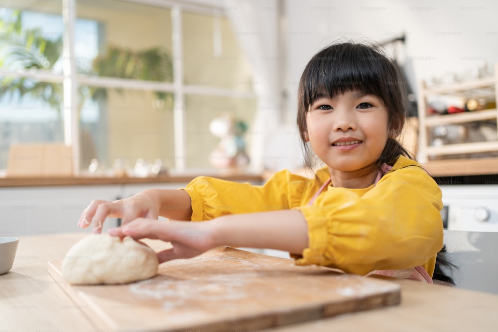 Retrato de una niña asiática haciendo panadería casera en la cocina. Adorable niño pequeño sentado en la mesa sintiéndose feliz y disfrutando aprender a cocinar alimentos u hornear amasa masa de levadura con las manos en casa