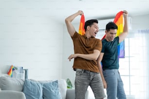 Asian handsome man gay family holding LGBTQ flag and dance together. Attractive romantic male lgbt couple spend time and enjoy listen to music, have fun with gay pride feeling and carry rainbow flag.