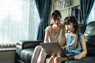 Giovane ragazza asiatica che impara a scuola a casa con la madre a casa. Il bambino piccolo guarda al computer e studia dall'insegnante di scuola dalla riunione digitale a distanza di Internet a causa della pandemia di coronavirus in casa