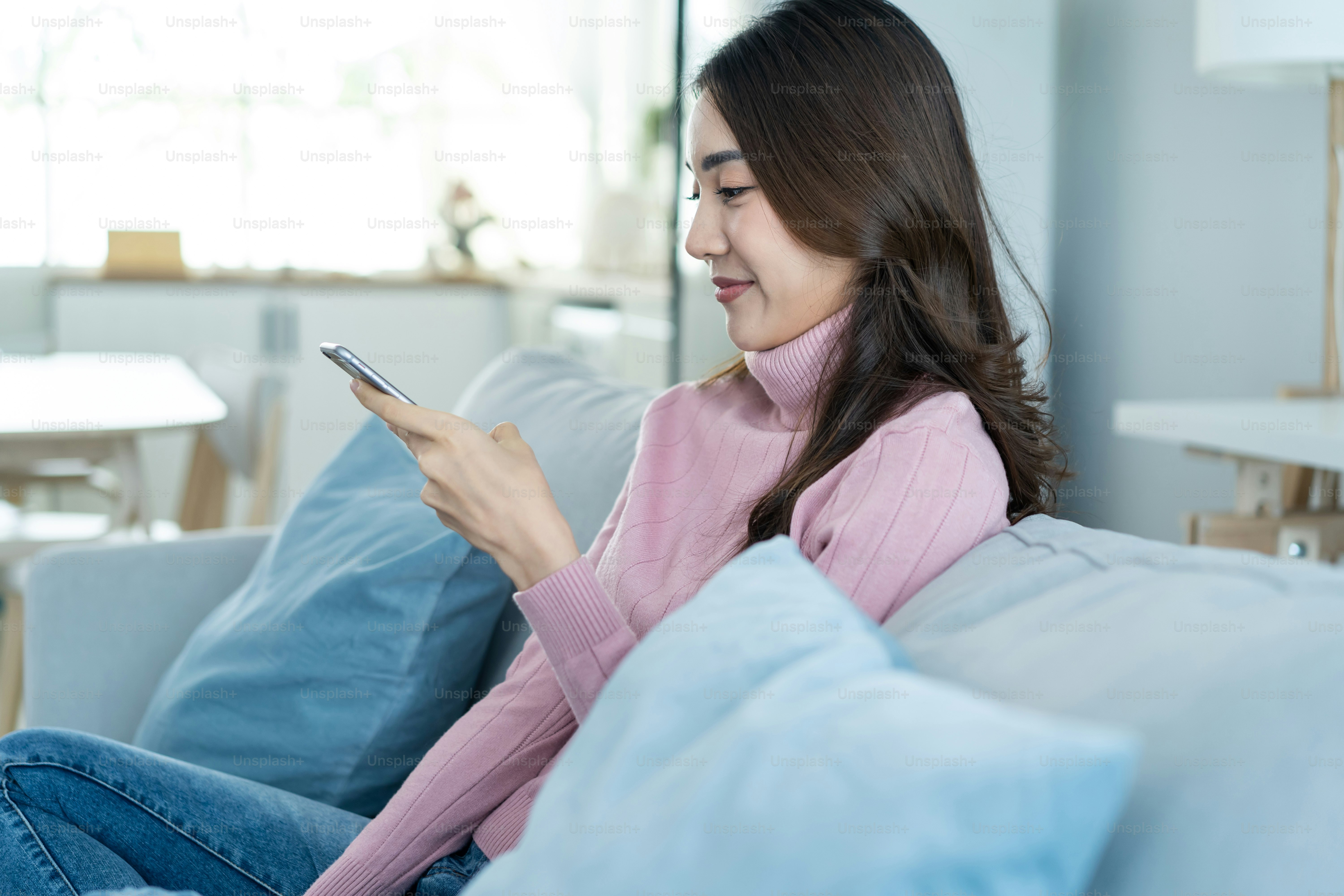 La bella donna asiatica si siede sul divano e chatta sul cellulare in casa. La giovane ragazza attraente felice trascorre il tempo libero a casa, si rilassa e si diverte a comunicare e discutere sullo smartphone in salotto.