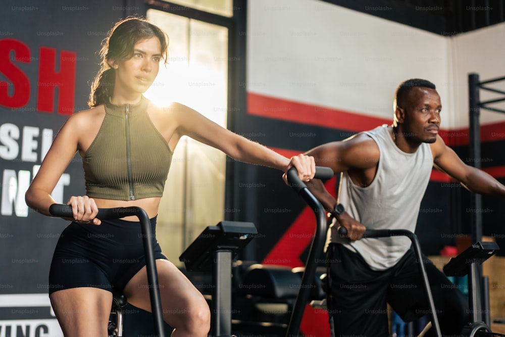 Caucasian athlete woman and African man exercise in fitness stadium. Attractive active sportsman and women workout together by cycling on bike machine for health care and wellbeing in modern gym club.