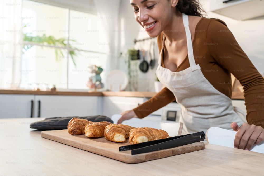 Mulher latina jovem atraente assando croissant na mesa na cozinha. Bonito avental feminino se sentir feliz e desfrutar de passar o tempo de lazer cozinhando padaria em casa. Atividade caseira em conceito de casa.