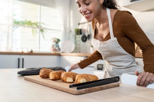 Attraente giovane donna latina che cuoce croissant sul tavolo in cucina. La bella donna indossa il grembiule si sente felice e si diverte a trascorrere il tempo libero cucinando panetteria a casa. Attività fatta in casa in casa.