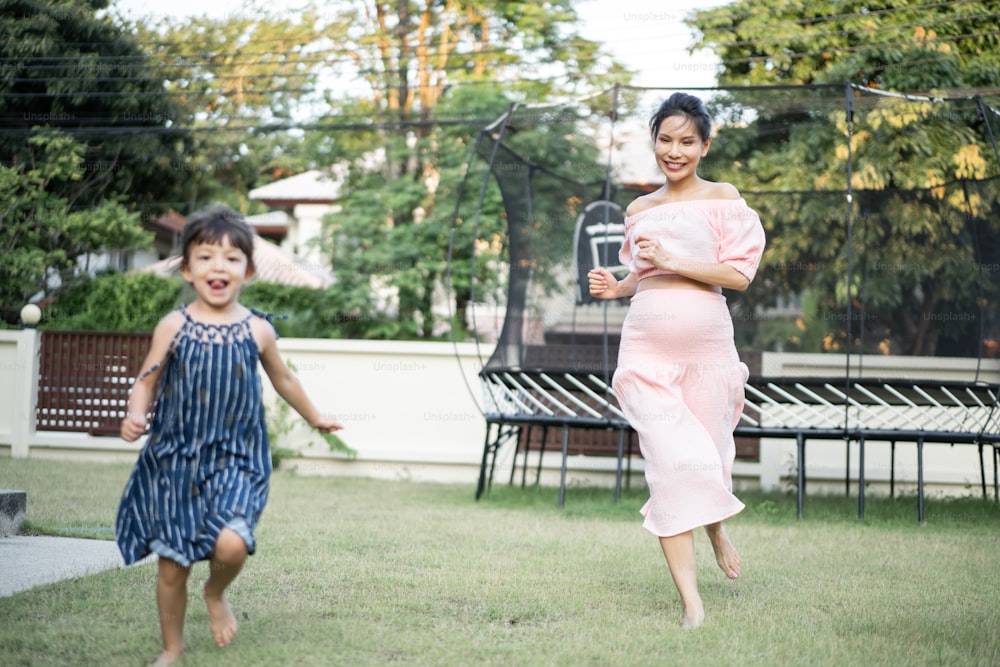 Asian young daughter and pregnant mother play together in garden home. Attractive beautiful pregnancy girl and little cute kid girl spending time running together with happiness and fun at backyard.
