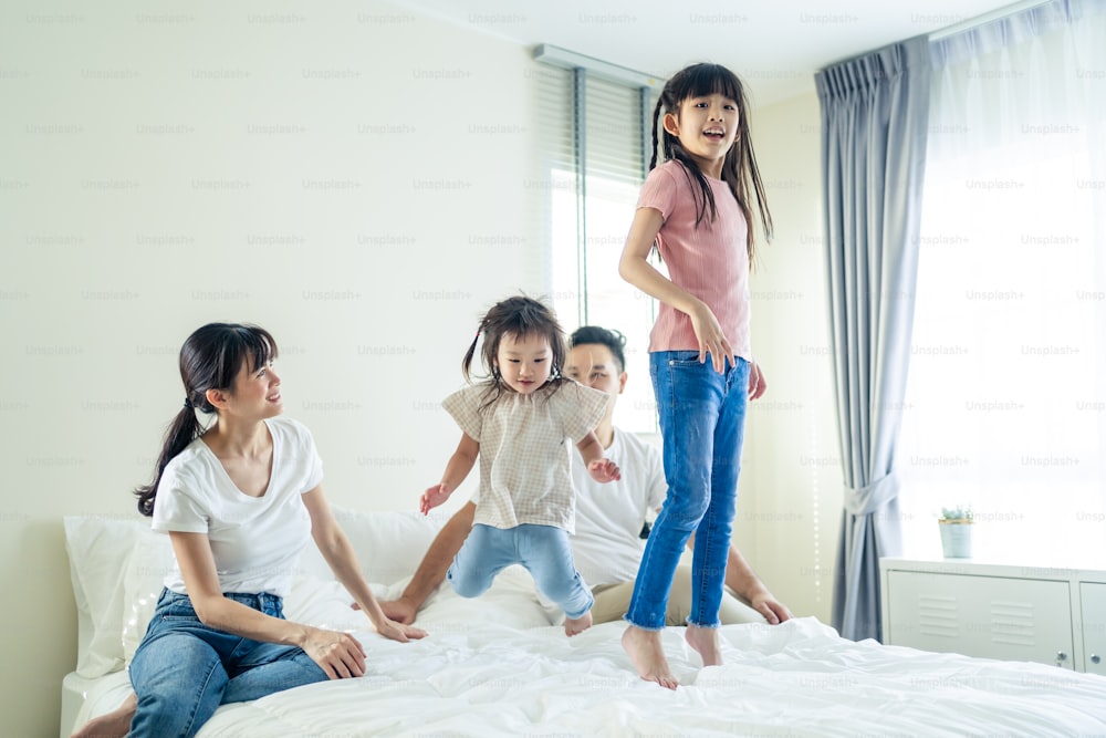 Asian funny family feel happy to spend time together on bed at home. Loving couple enjoy look to young little girl daughters playing and jumping on bed in the morning together after wake up at house.