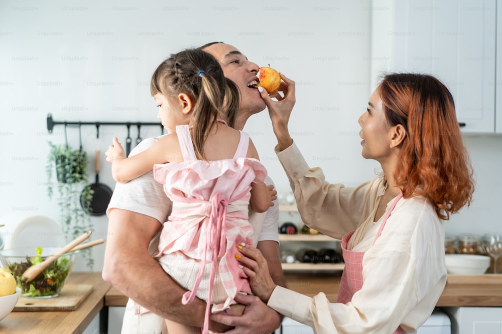 Asian attractive parents spend time with cute baby daughter in kitchen. Happy family, loving couple mother feed apple to husband while holding little kid girl on hands in house. Activity relationship