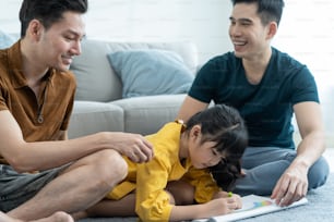 Família gay LGBTQ atraente asiática ensinar jovem menina desenhar imagem. Casal masculino bonito olhar para a criança pouco adorável filha brincar e livro de colorir na sala de estar, desfrutar de atividade parental em casa