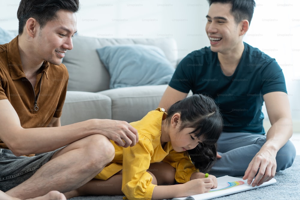 Asiatische attraktive LGBTQ schwule Familie lehren junges Mädchen Kind zeichnen Bild. Ein hübsches männliches Paar schaut sich das kleine entzückende Kind an, spielt und malt Buch im Wohnzimmer, genießt die elterliche Aktivität zu Hause