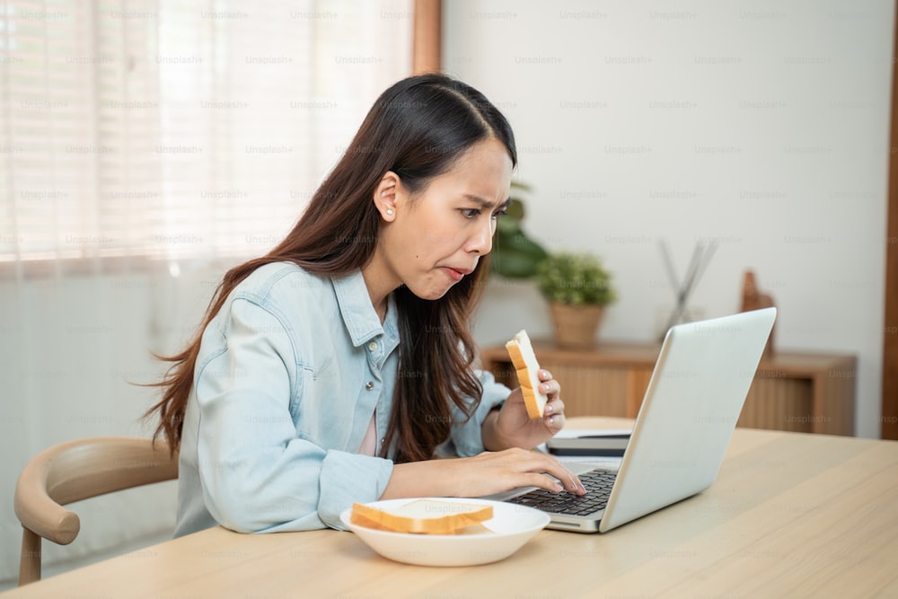 Asiatische attraktive Geschäftsfrau, die hungrig Brot in Eile isst, während sie Geschäfte auf Notebook-Computer macht. Schöne ernsthafte Studentin, die sich frustriert und beschäftigt fühlt, zu Hause zu arbeiten und zu lernen